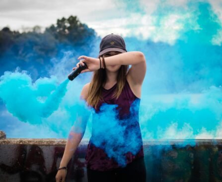 A stylish woman surrounded by bright blue smoke outdoors, creating a dynamic fashion statement.